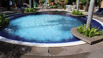 A swimming pool with a fountain, surrounded by lounge chairs and tables, with a building in the background.