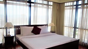 Una cama grande con sábanas blancas y un cabecero oscuro, flanqueada por dos mesitas de noche con lámparas, en una habitación con ventanales y cortinas translúcidas.