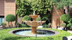 Una fuente circular con agua que cae en varios niveles, rodeada de setos cuidados y plantas en macetas.
