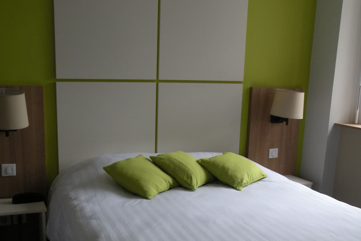 A neatly made bed with white bedding and three green pillows, a wooden nightstand with a lamp, and a green wall with a large white panel above the bed.