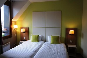 Hotellihuone, jossa on kaksi sänkyä, molemmissa valkoiset vuodevaatteet ja vihreät tyynyt, suuri ikkuna ja seinään kiinnitetyt lamput.