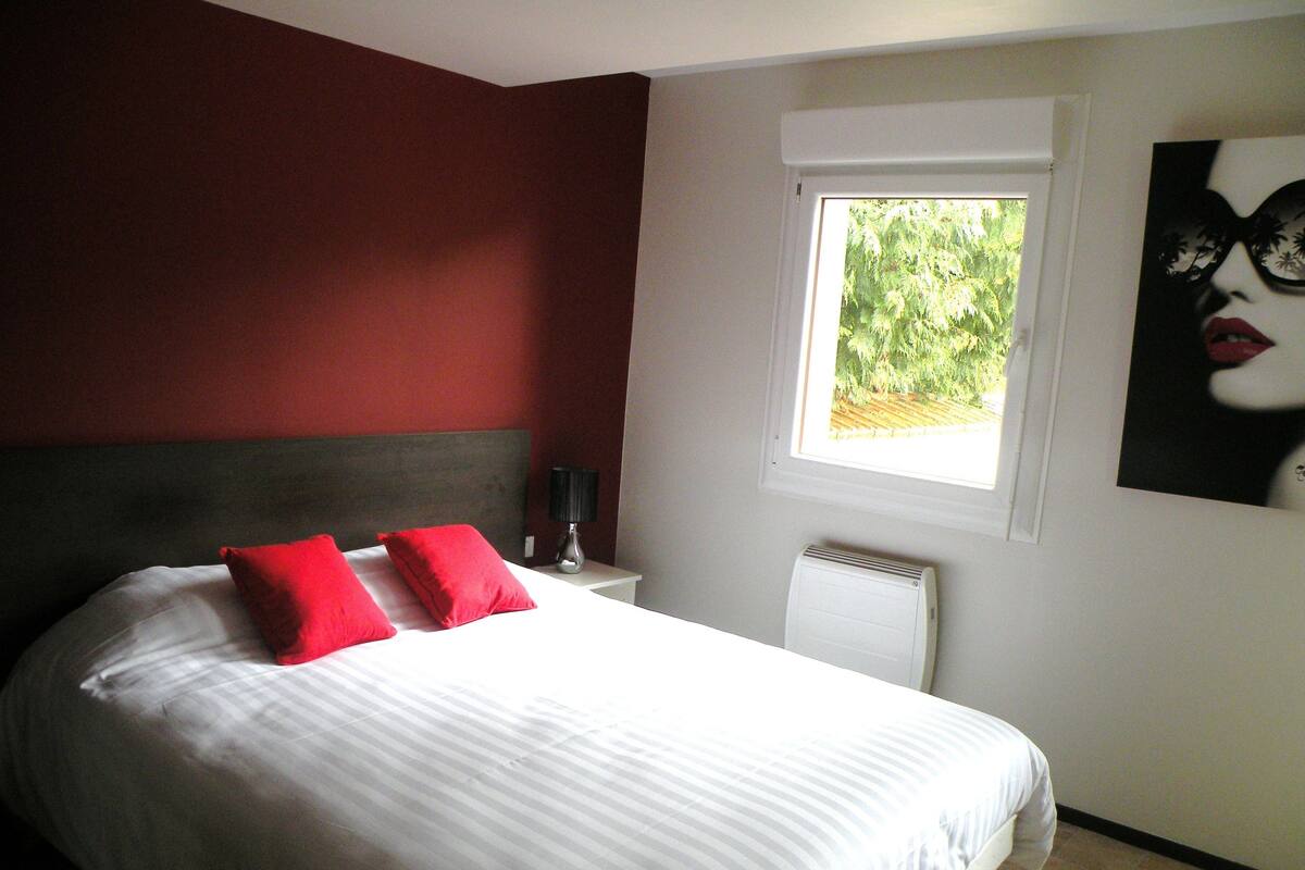 A bedroom with a bed, red pillows, a nightstand, a lamp, a radiator, and a window with a view of greenery.
