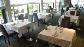 Un restaurante con varias mesas dispuestas para comer, que cuenta con una alfombra estampada y amplios ventanales que dejan entrar la luz natural.