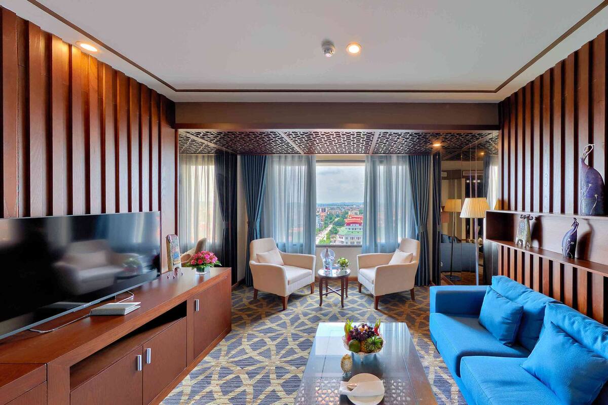 A hotel room with a TV, sofa, two chairs, a coffee table, and a view of the city.