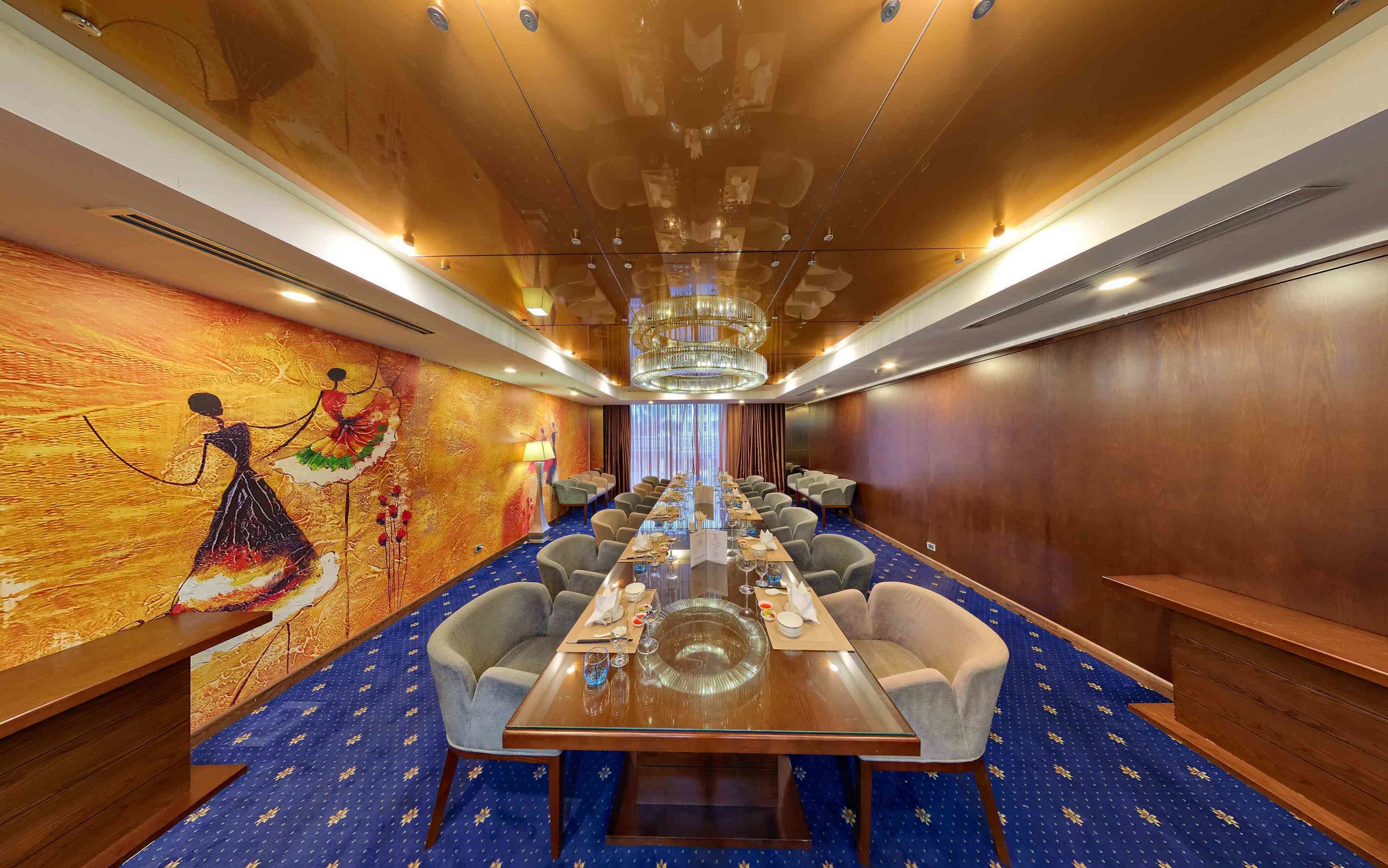 Un salón de conferencias con una larga mesa dispuesta para una reunión, rodeado de sillas, con un mural en la pared y una araña colgante.