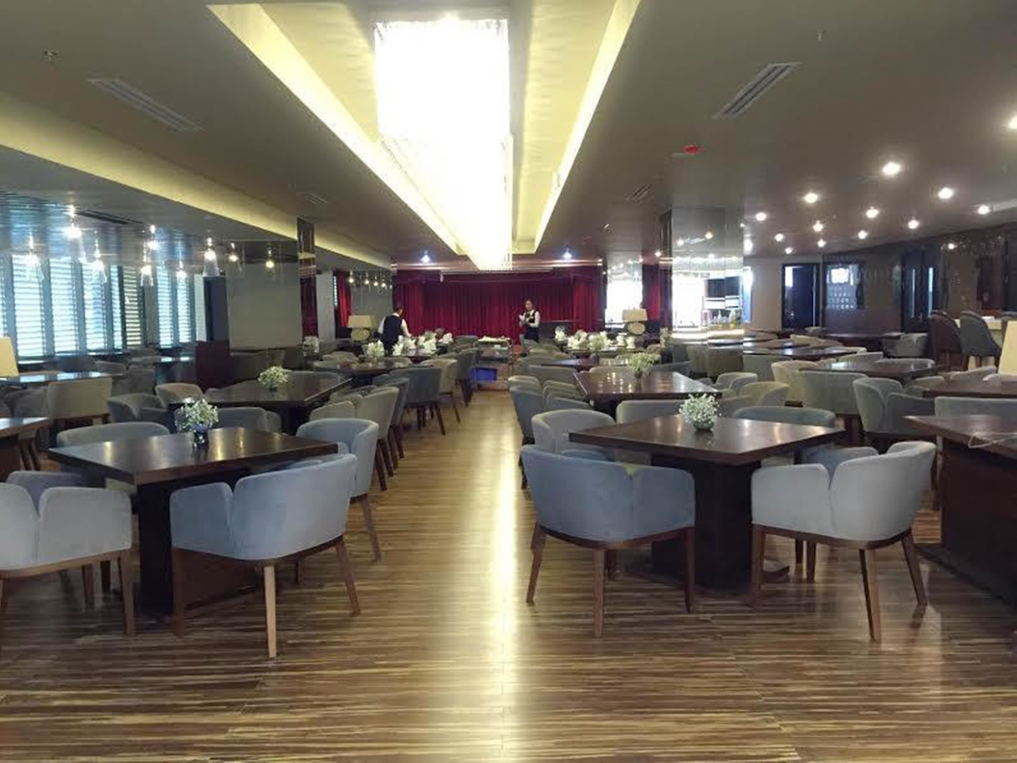 A spacious dining area with multiple tables and chairs, a stage at the far end, and large windows along one wall.