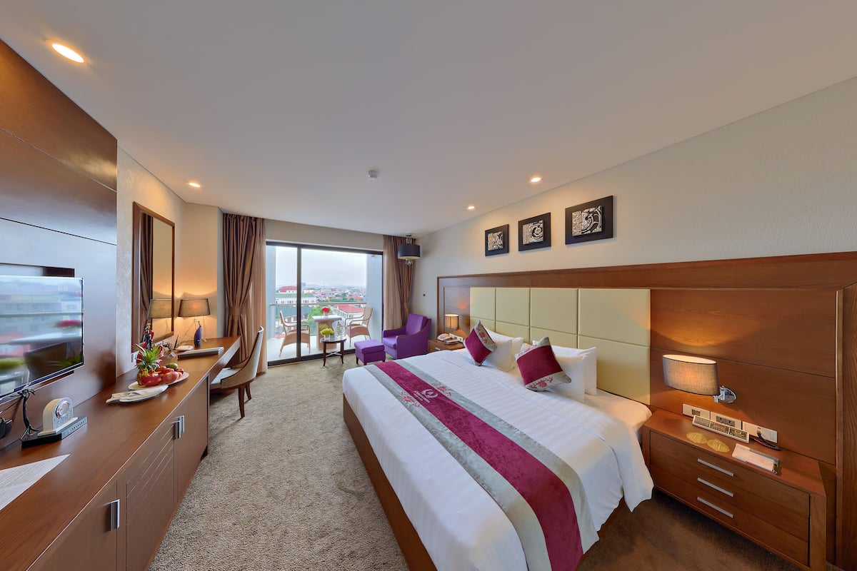 A hotel room with a large bed, a desk with a flat-screen TV, a chair, and a view of the city.