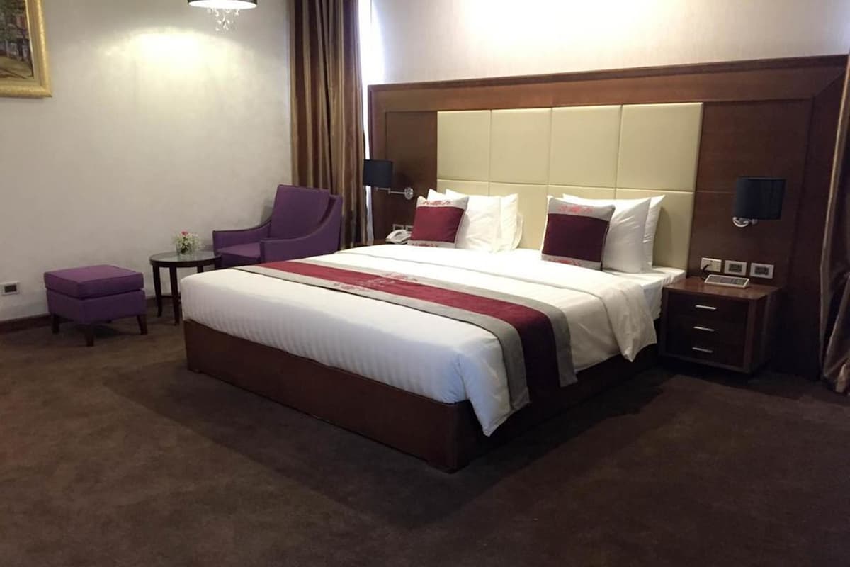 A hotel room with a large bed, a chair, a small table, and a nightstand with a phone and a lamp.