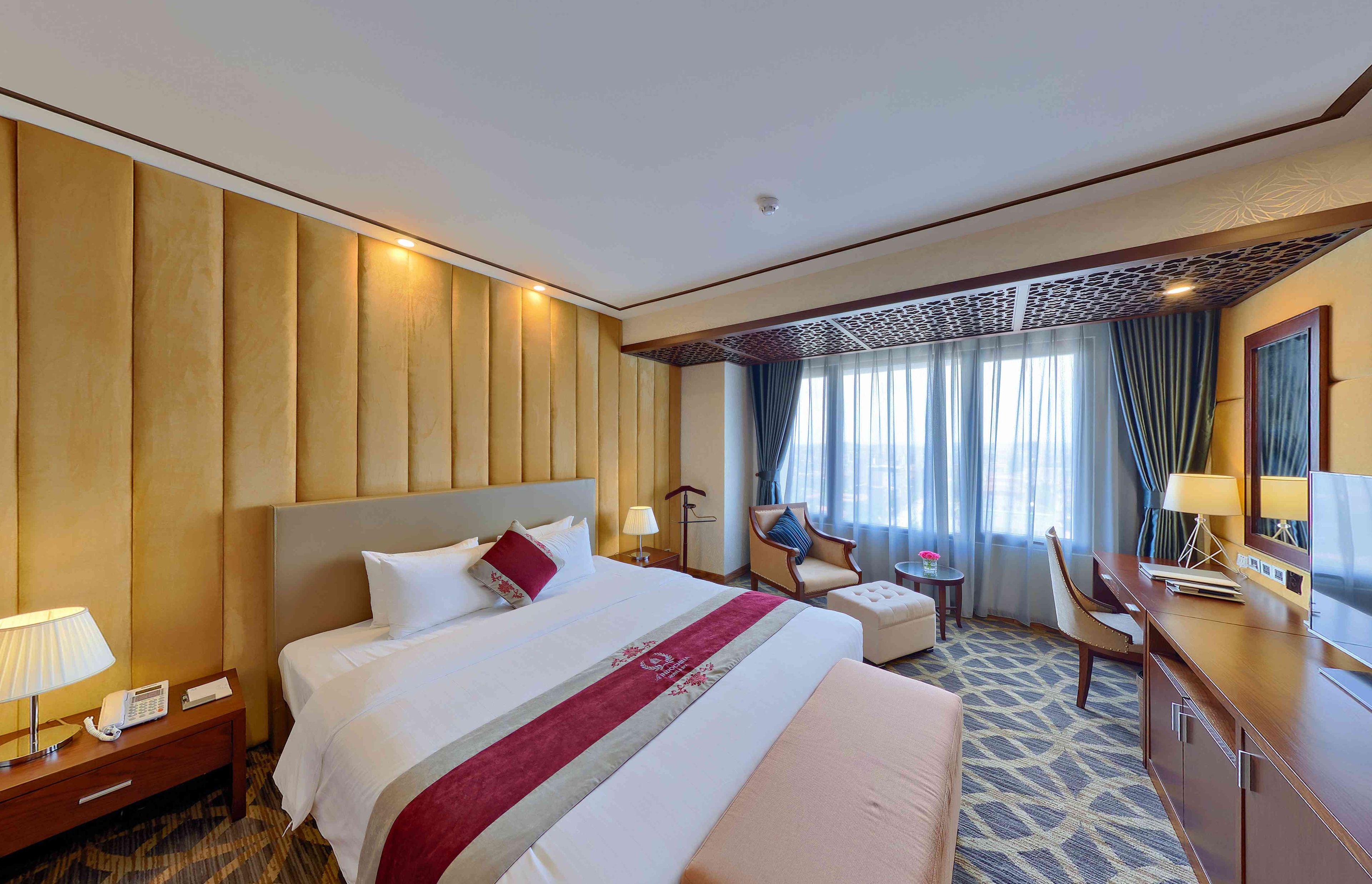 Ein Hotelzimmer mit einem großen Bett, einem Schreibtisch, einem Sessel, einem Nachttisch mit Telefon und einem Fenster mit Vorhängen.
