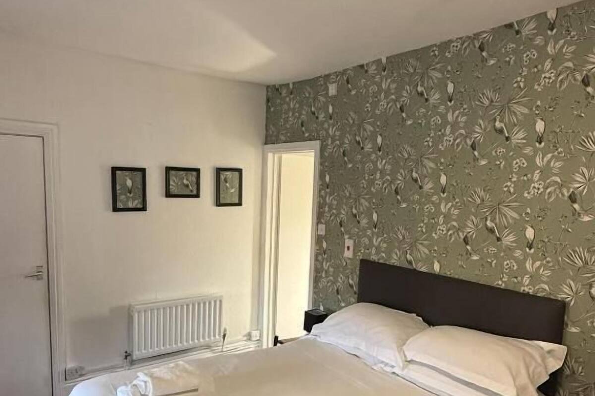 A neatly made bed with white linens and towels, a radiator, a door, and a wall with a floral pattern.