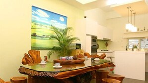 A modern dining area with a unique wooden table and stools, a painting on the wall, and a kitchen in the background. - Langkawi Garden B&B (Luodong)