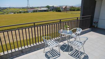 A balcony with two metal chairs and a small table, overlooking a grassy field and buildings in the distance.