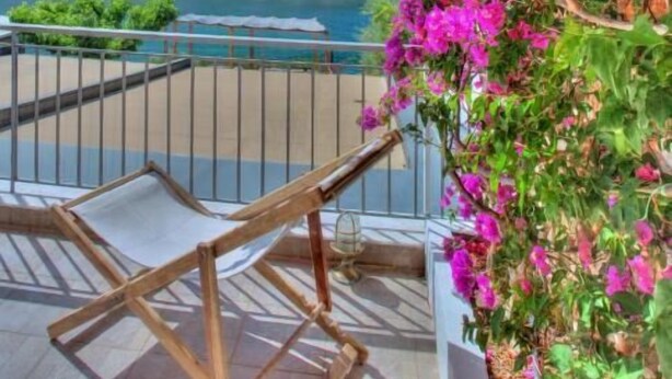 Un balcón con vistas a un lago y montañas, sillas de madera y flores de buganvilla rosadas.