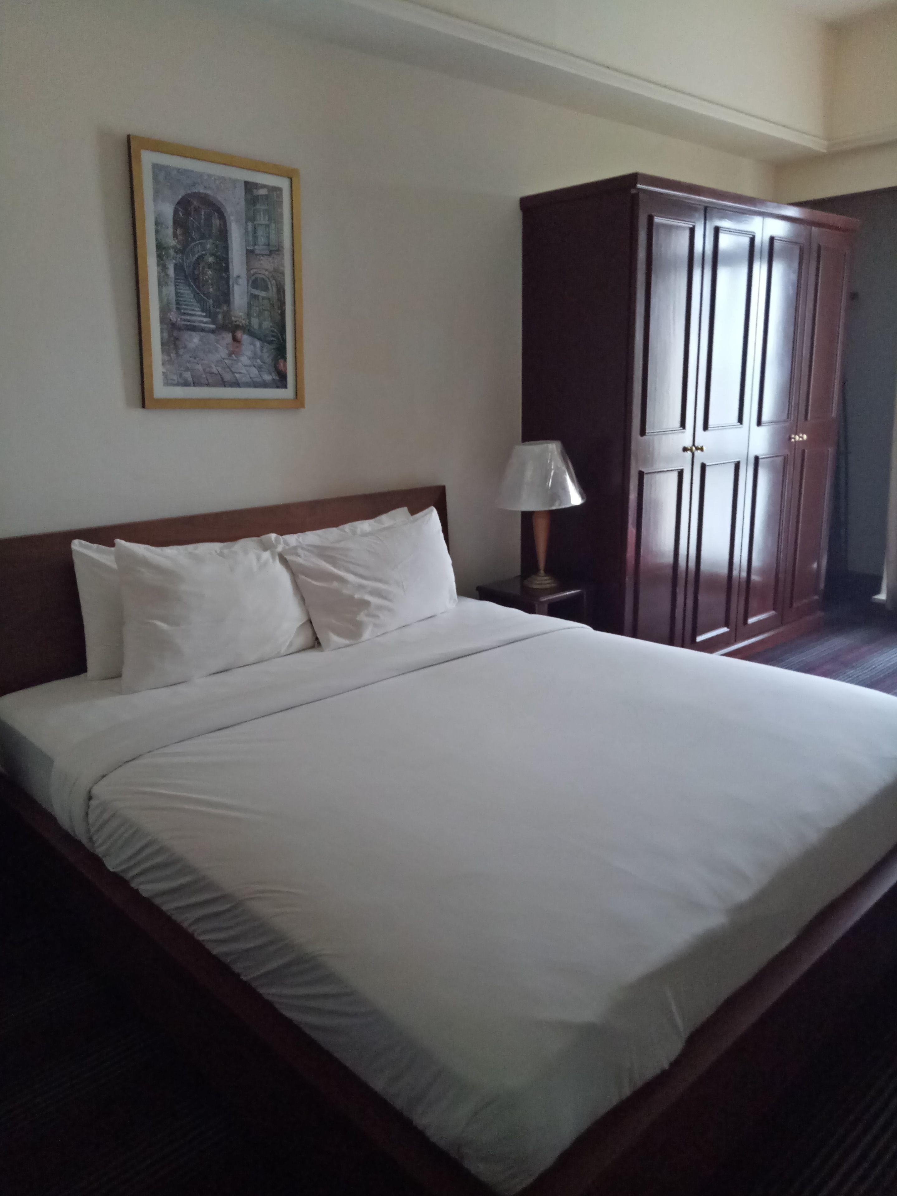 A neatly made bed with white linens, a wooden headboard, and a nightstand with a lamp. A framed picture hangs on the wall above the bed.