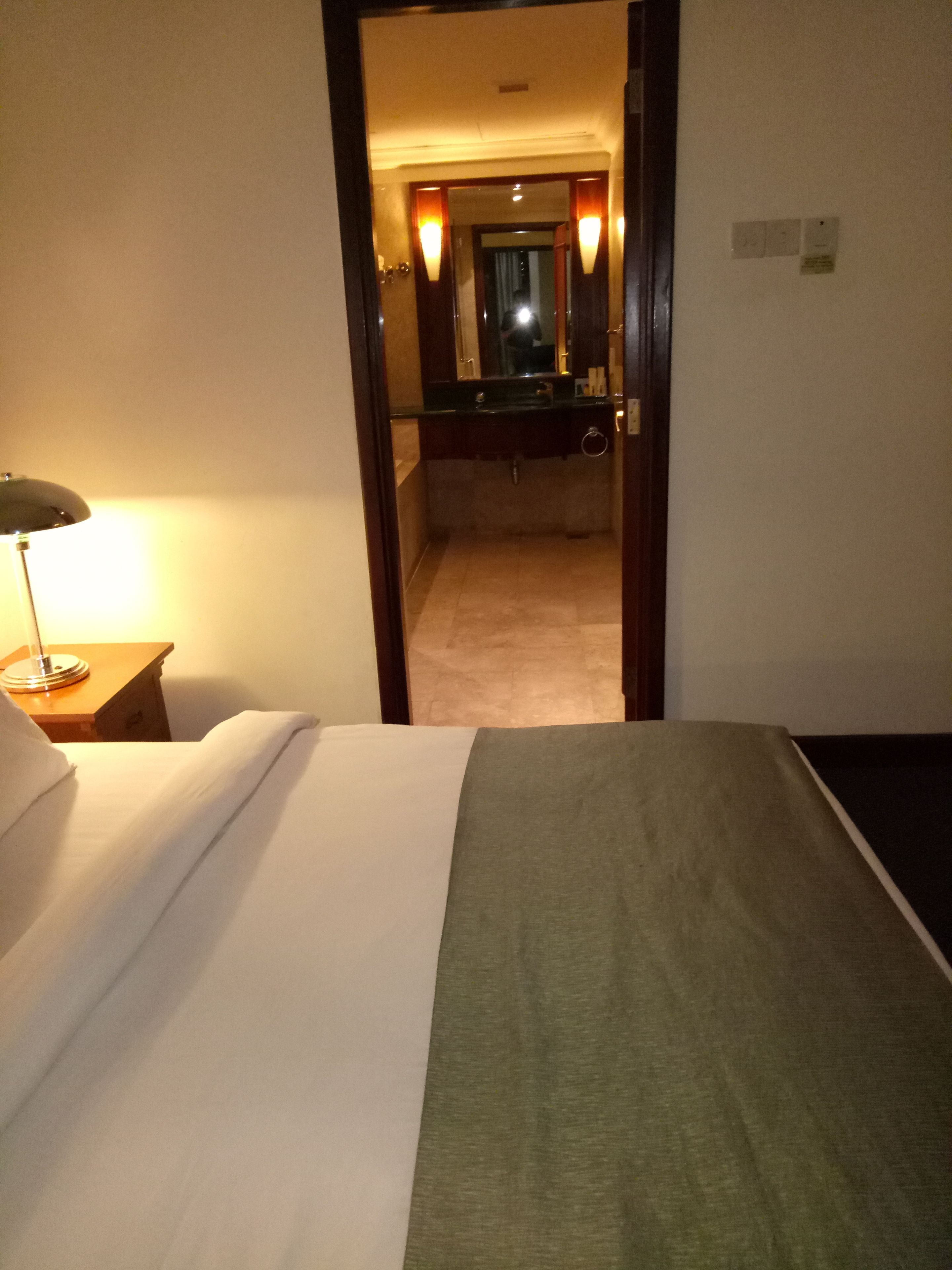 A hotel room with a bed, a bedside lamp, a bathroom visible through an open door, and a mirror with lights.