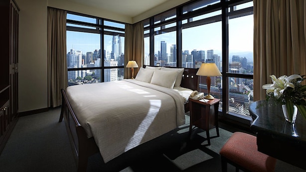 A hotel room with a large bed, two bedside lamps, a desk with a telephone, a vase with flowers, and a city view through large windows. - Sky Apartment at Times Square (Kuala Lumpur)
