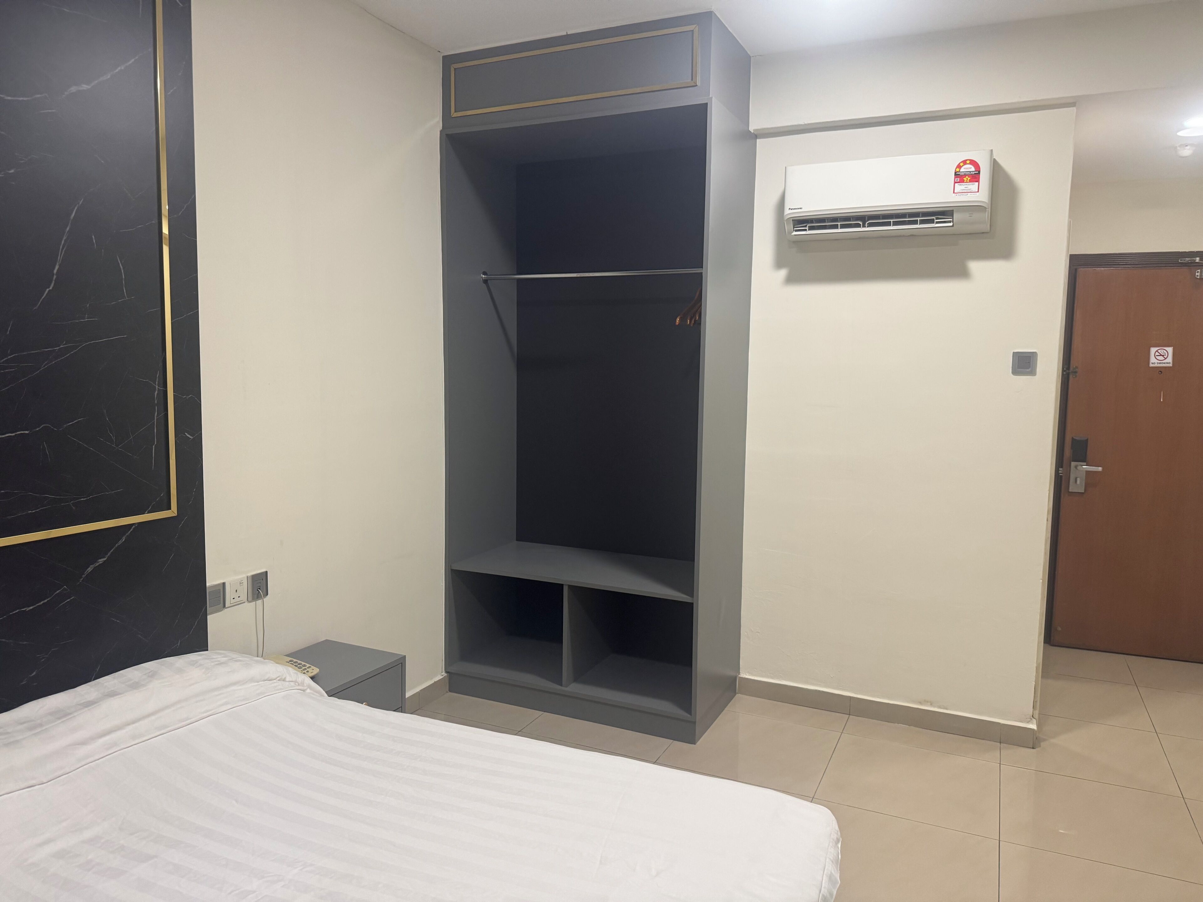 A hotel room with a bed, a wardrobe, a bedside table, an air conditioning unit, and a door.