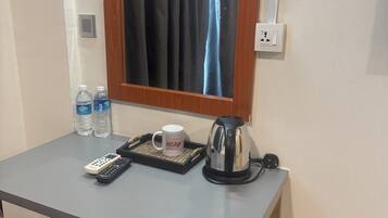 Una habitación de hotel pequeña con un escritorio, una silla, un hervidor, una bandeja con una taza, dos botellas de agua y un control remoto.
