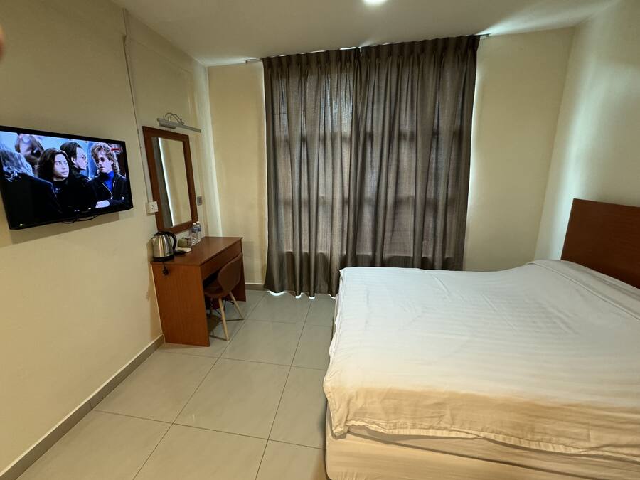 A hotel room with a bed, a desk with a chair, a television mounted on the wall, and a large window with curtains.