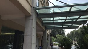 A building with a glass-covered entrance, a concrete pillar, and a view of a street with parked cars and greenery. - Aeton Hotel (Nilai)