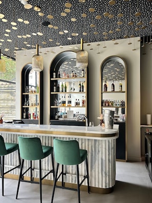 Een bar met een marmeren aanrecht, groene barkrukken en planken vol flessen.