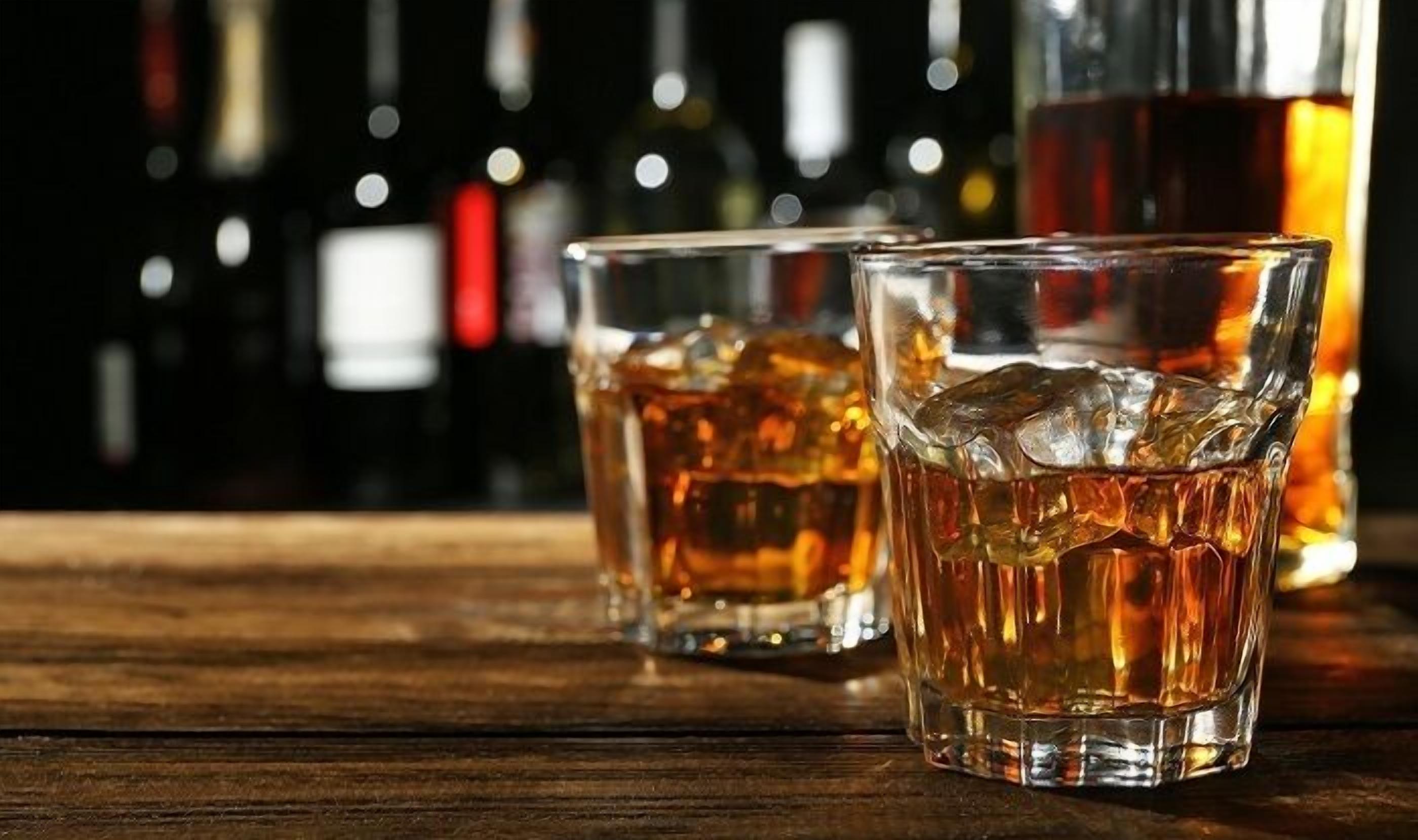 Two glasses of whiskey on a wooden surface with bottles in the background.