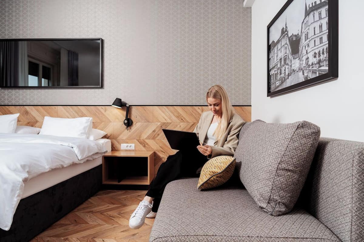 Habitación de hotel con cama, sofá, televisión montada en la pared, mesita de noche y un cuadro enmarcado.