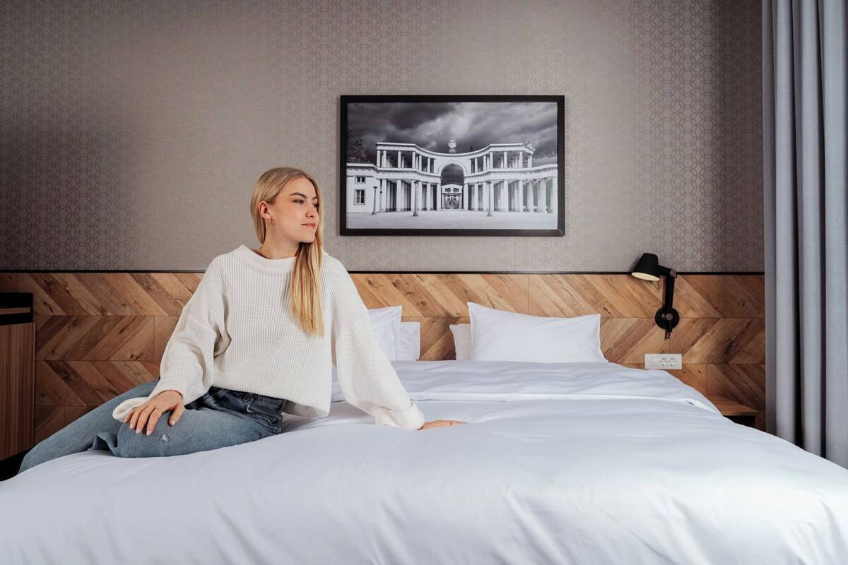 Una persona sentada en una cama con cabecera de madera, un cuadro enmarcado en la pared y una lámpara de noche.