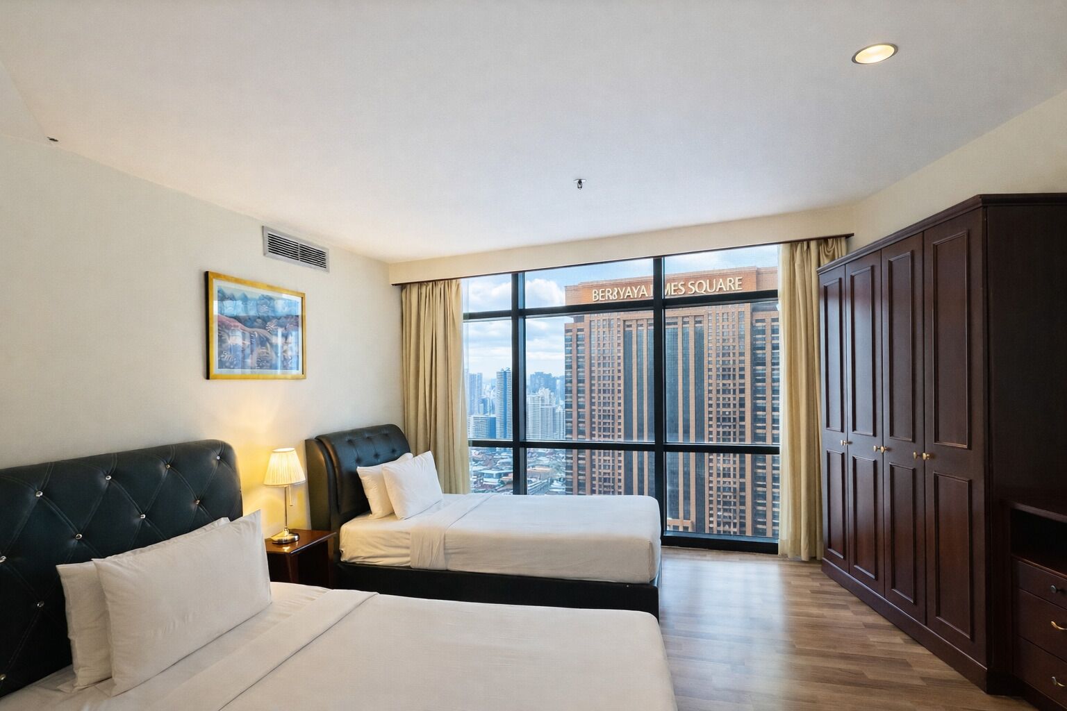 A hotel room with two beds, a large window with a city view, a dark wooden wardrobe, and a framed picture on the wall.