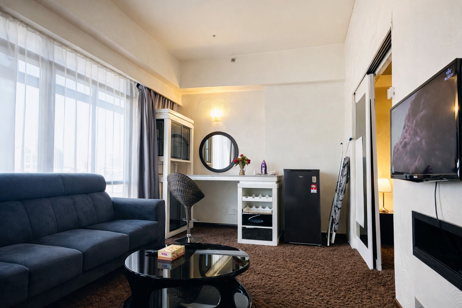 A hotel room with a sofa, a small table, a TV, a minibar, and a large window.