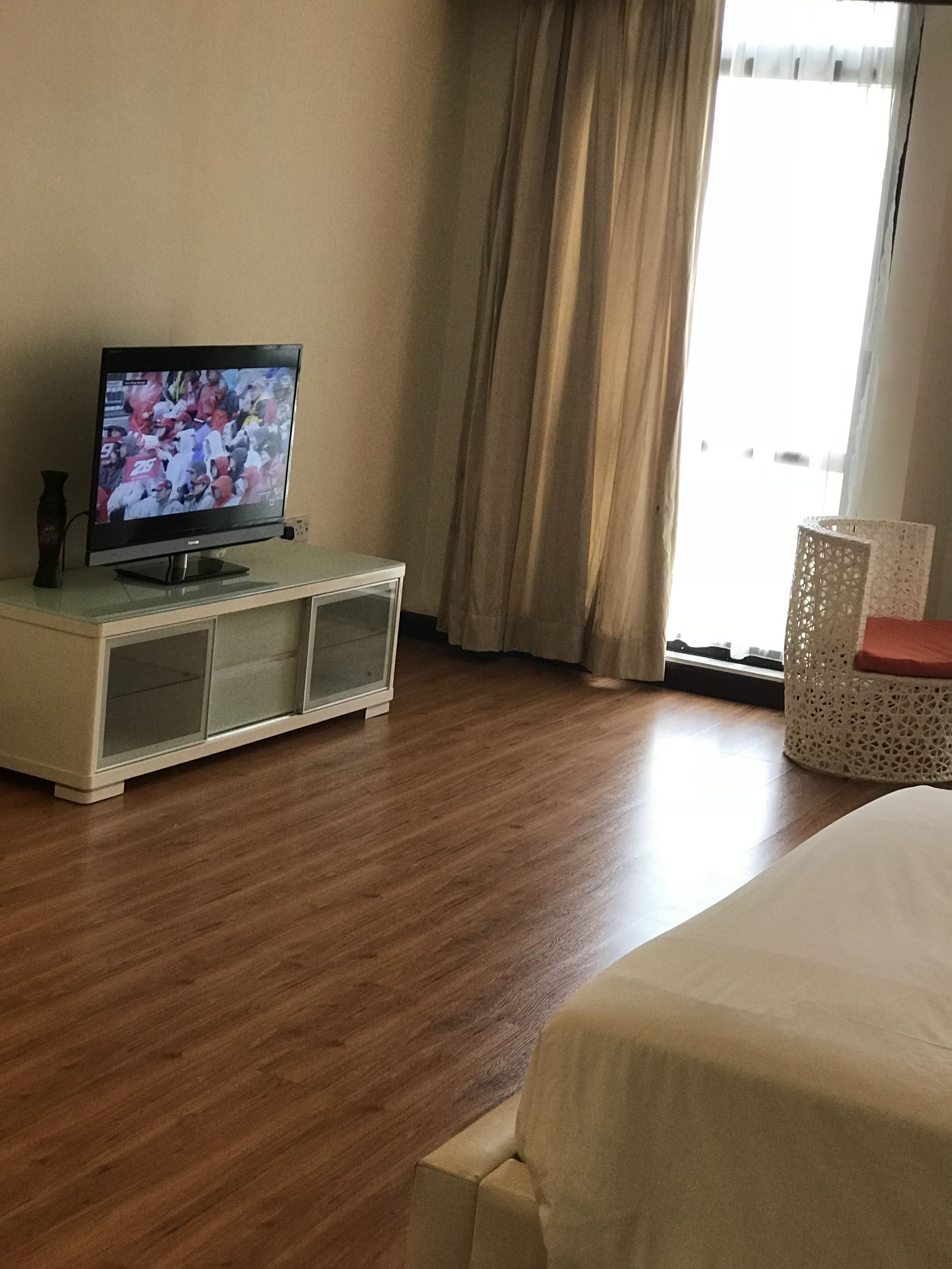 A hotel room with a television on a cabinet, a bed, a window with curtains, and a wicker chair.