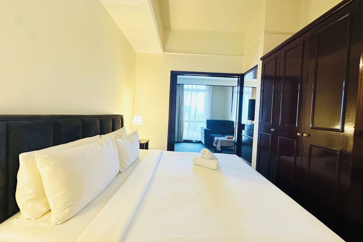 Ein Hotelzimmer mit einem großen Bett, einem Nachttisch, einer Lampe, einem Kleiderschrank und einer offenen Tür, die in einen anderen Raum führt.