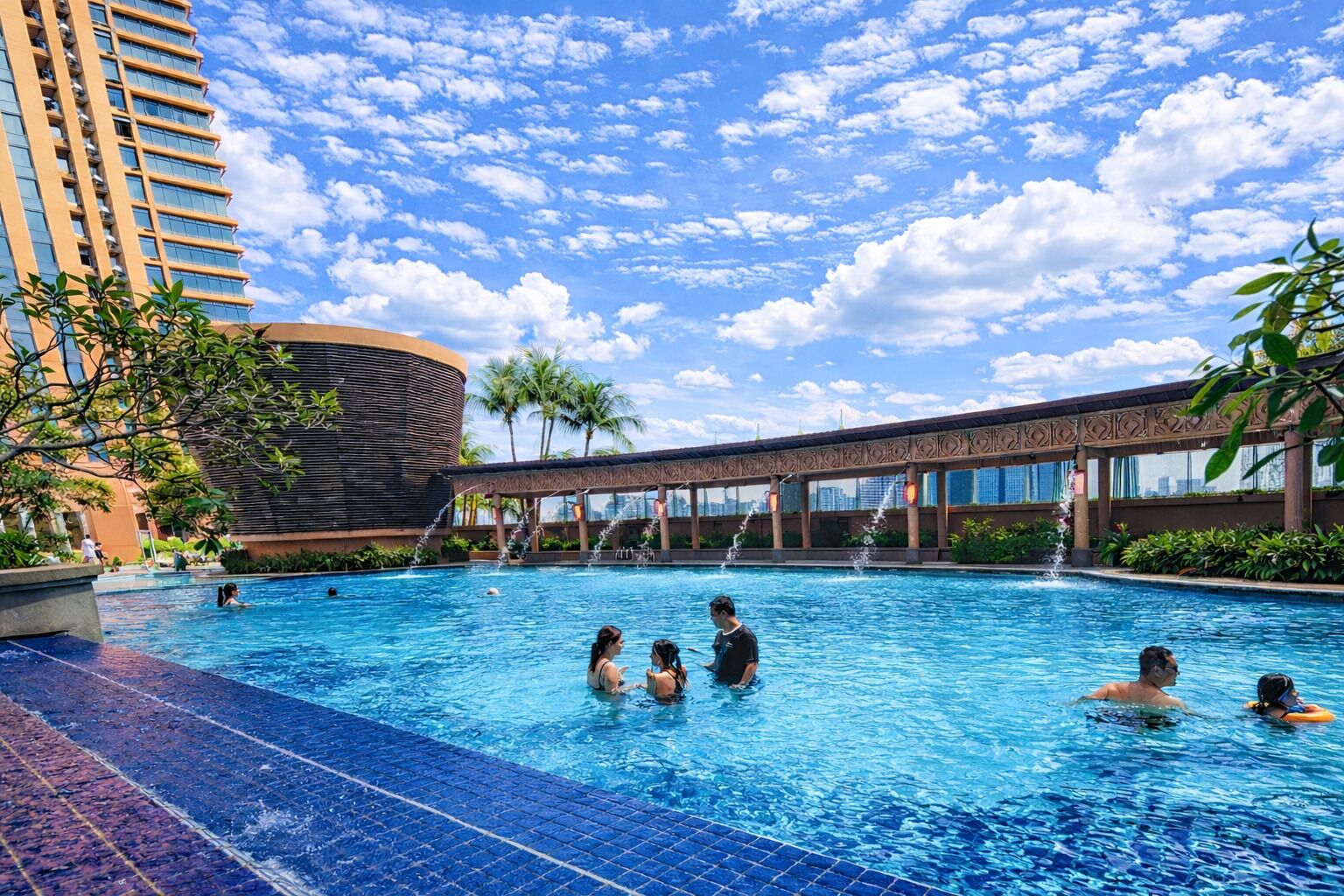 A swimming pool with people, a building with a curved facade, and a bridge with water features.
