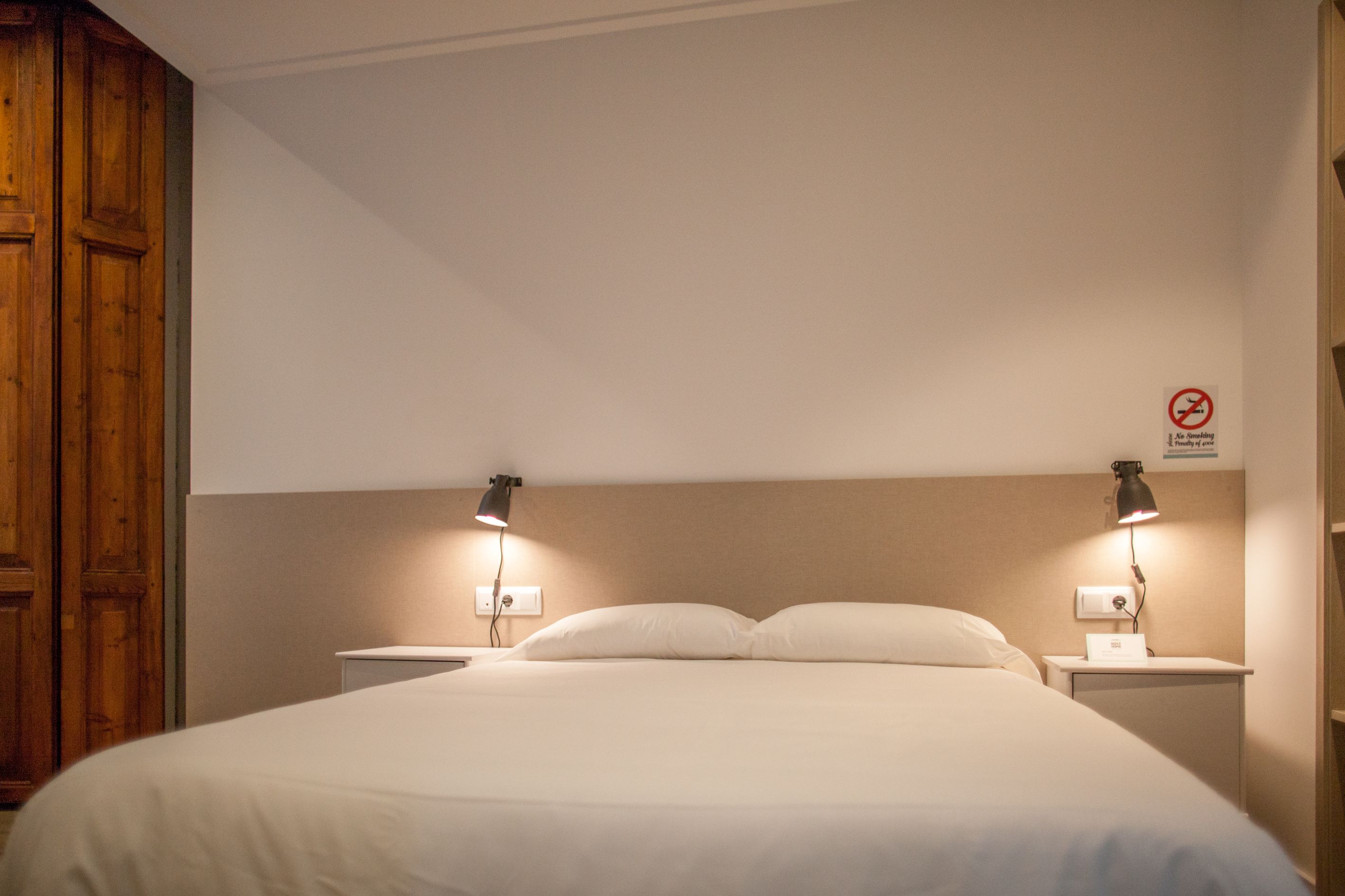 A hotel room with a large bed, two bedside lamps, a wooden wardrobe, and a no-smoking sign on the wall.