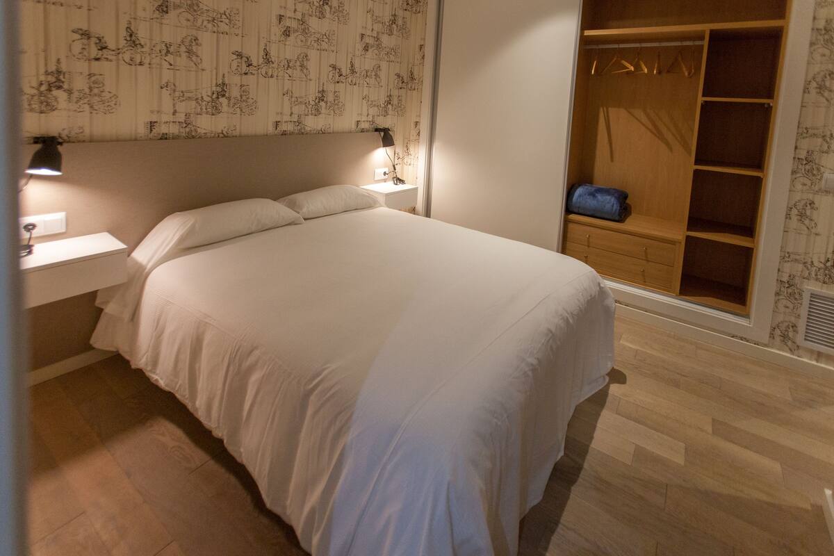 A neatly made bed with a headboard, a bedside table with a lamp, and a wooden wardrobe with hanging space.