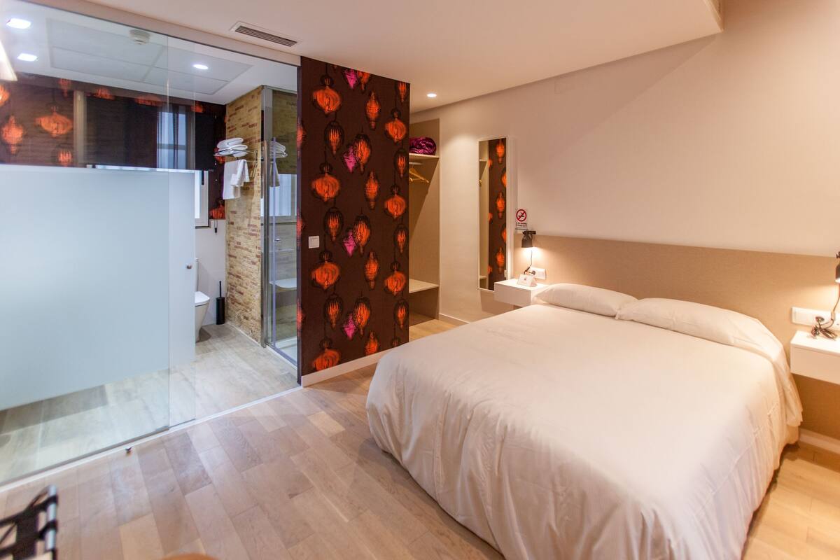 A modern hotel room with a large bed, a bathroom with a glass partition, and a small sitting area.