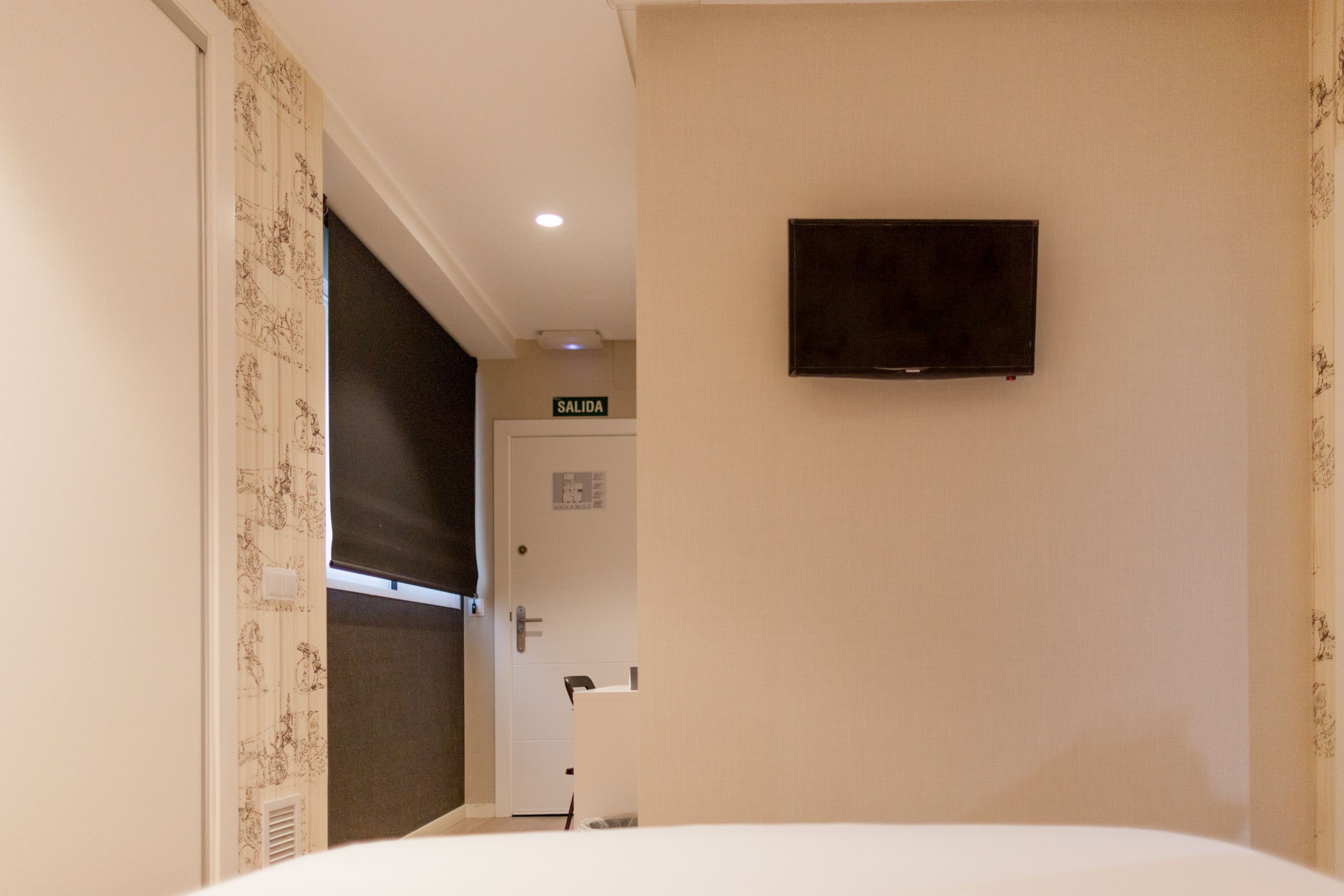 Habitación de hotel con cama, televisor montado en la pared, puerta con la leyenda 'SALIDA' y un respiradero en la pared.