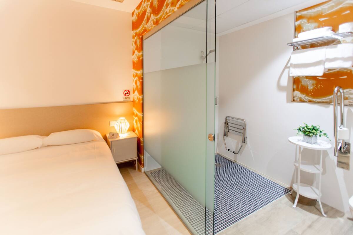 A hotel room with a bed, a nightstand, a glass shower enclosure, a small white table with a plant, and a towel rack.