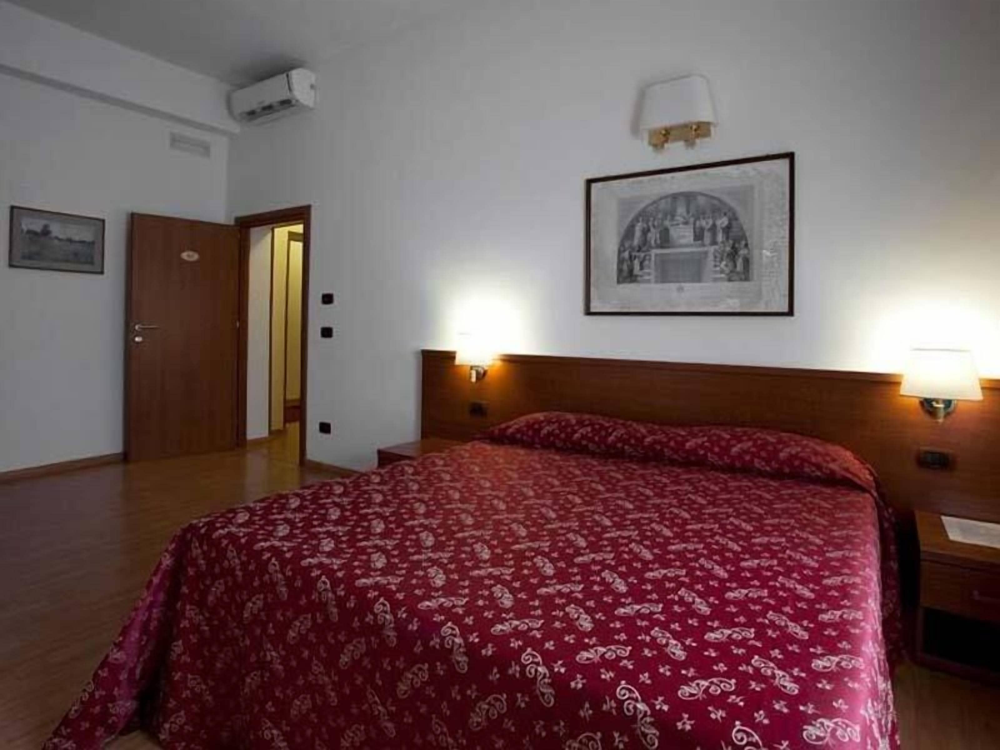 Quarto de hotel com uma cama grande, duas luminárias de cabeceira, um quadro na parede e uma porta que dá para outro quarto.