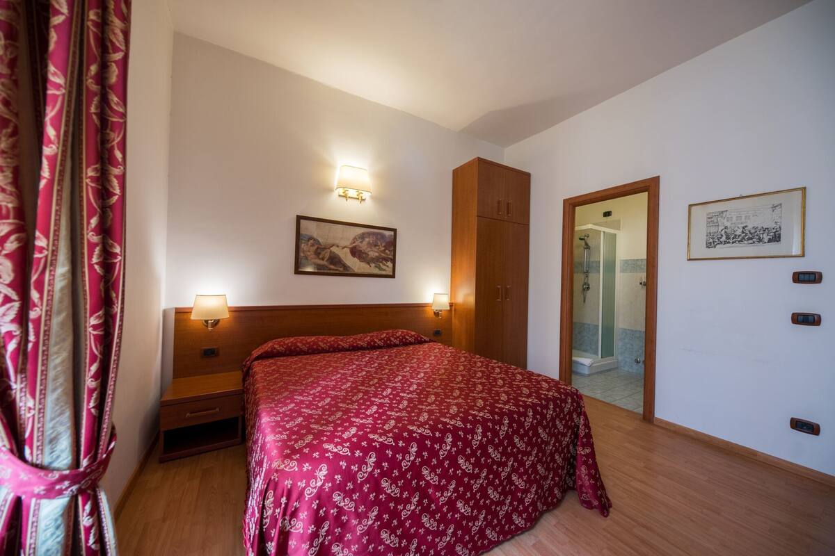 A bedroom with a wooden bed, two bedside lamps, a wooden wardrobe, a framed picture on the wall, and a door leading to a bathroom.