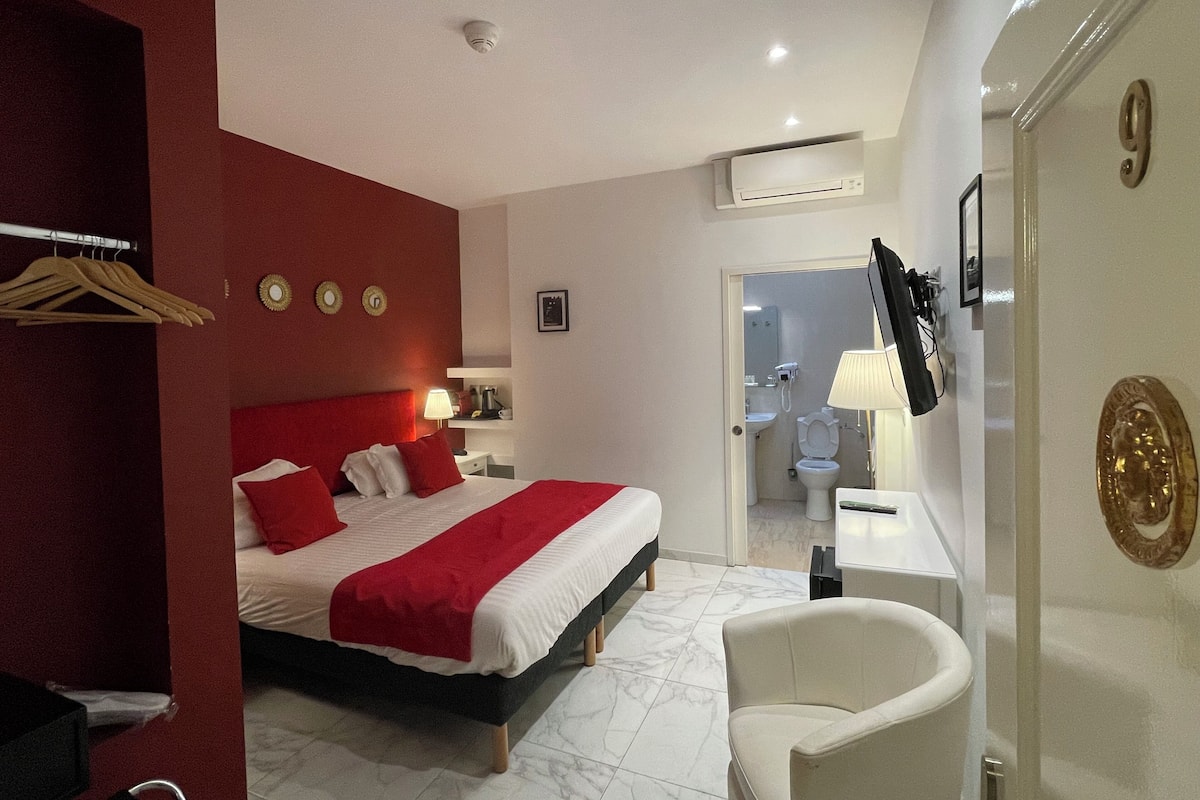A hotel room with a bed, a chair, a desk, and a bathroom visible through an open door.