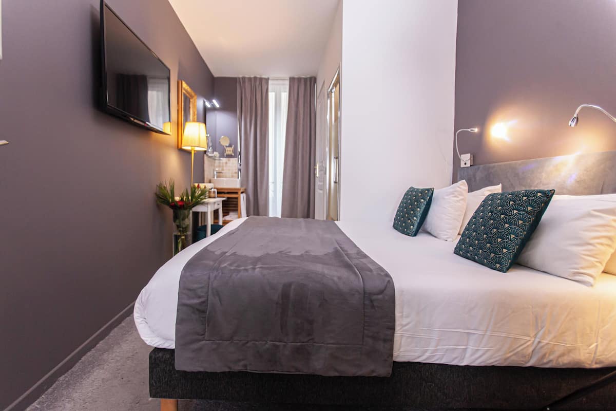 A hotel room with a large bed, a flat-screen TV mounted on the wall, a bedside table with a lamp, and a view of the hallway.
