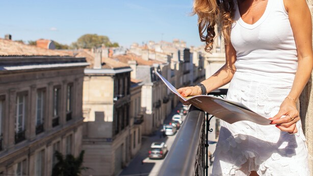 A person in a white dress standing on a balcony overlooking a city street with buildings and parked cars. - Hôtel Churchill (Bordeaux)