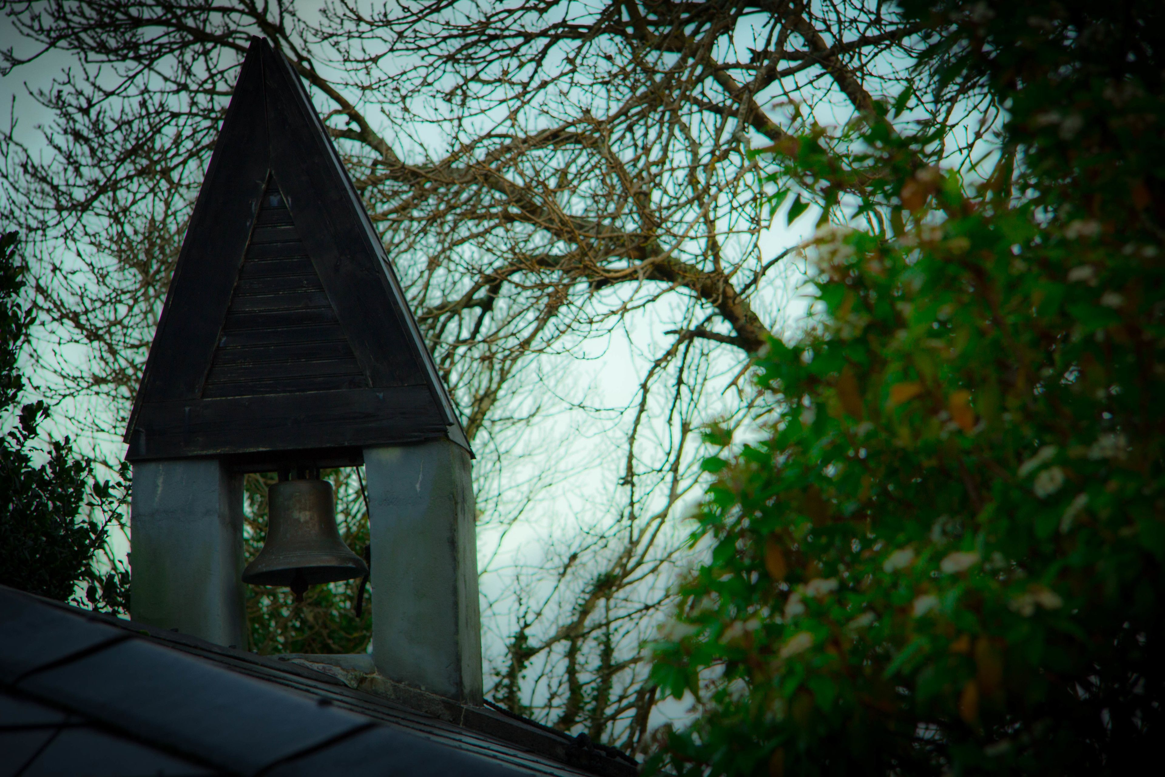 Un campanario con techo triangular y una campana colgando, rodeado de árboles.