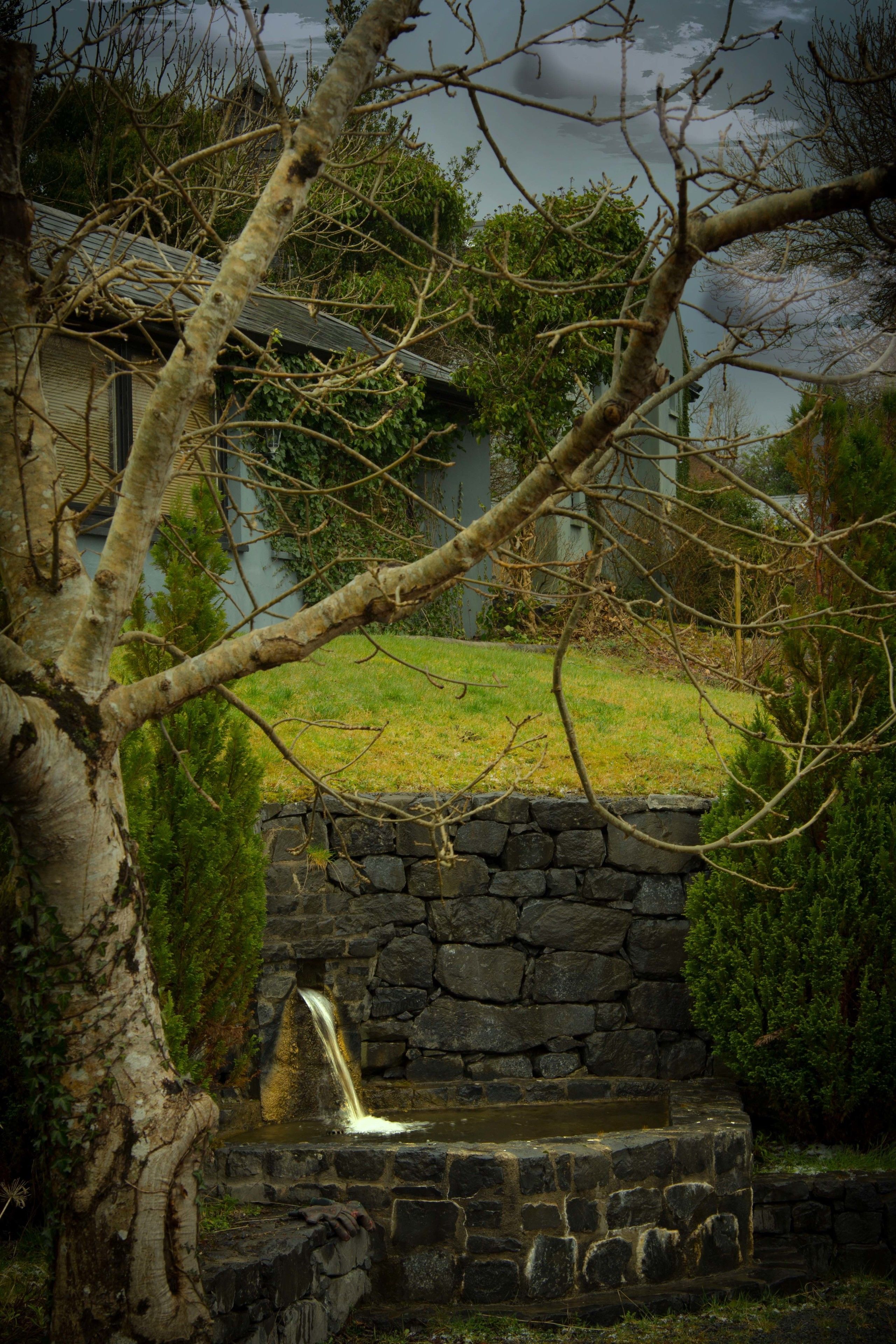 A stone water feature with a small waterfall, surrounded by a stone wall and a tree.