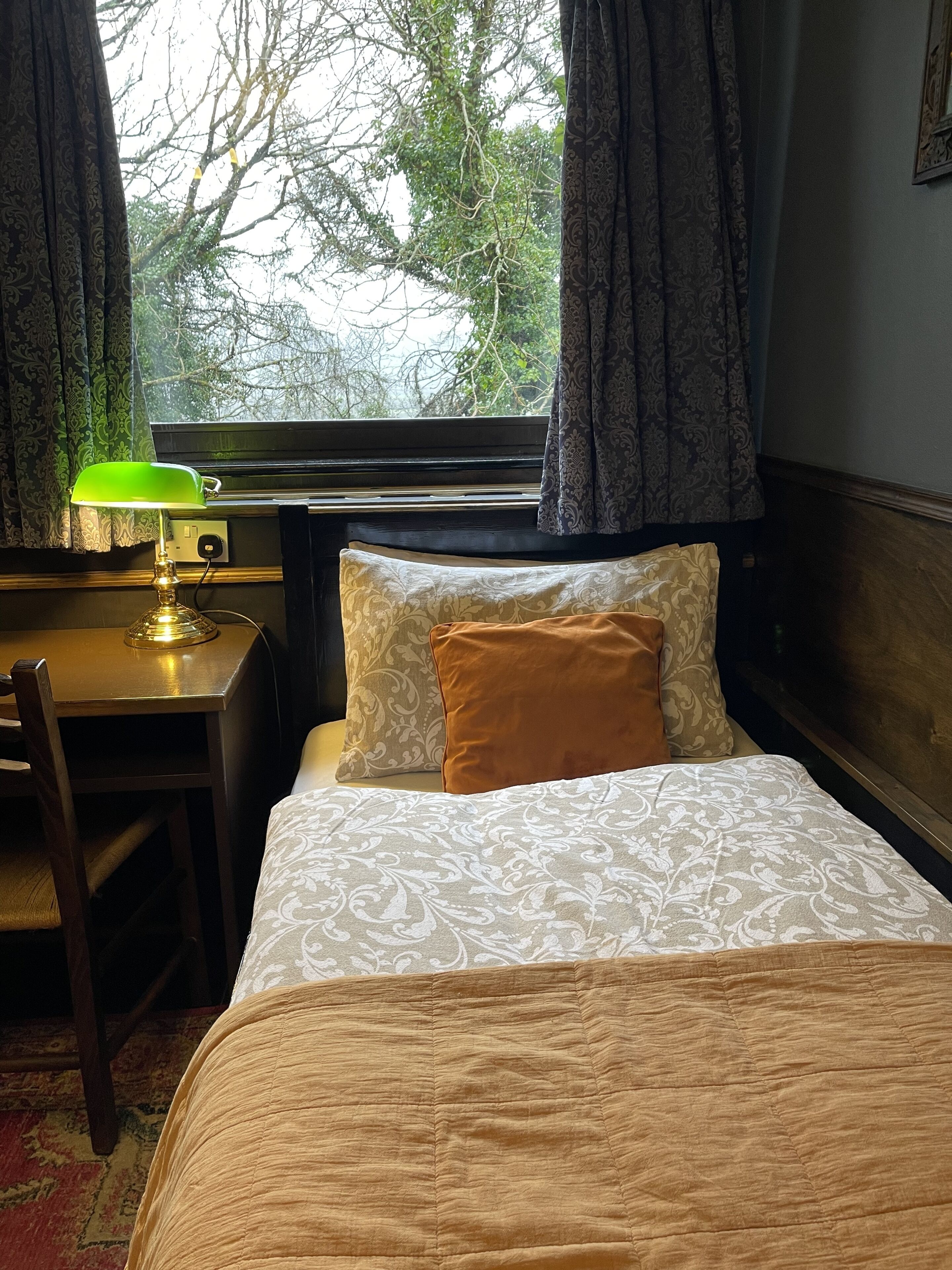 Habitación de hotel con cama, escritorio, lámpara y vistas a árboles por la ventana.