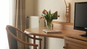 Una habitación de hotel con un escritorio de madera, una silla, un florero con flores, una lámpara y un televisor.