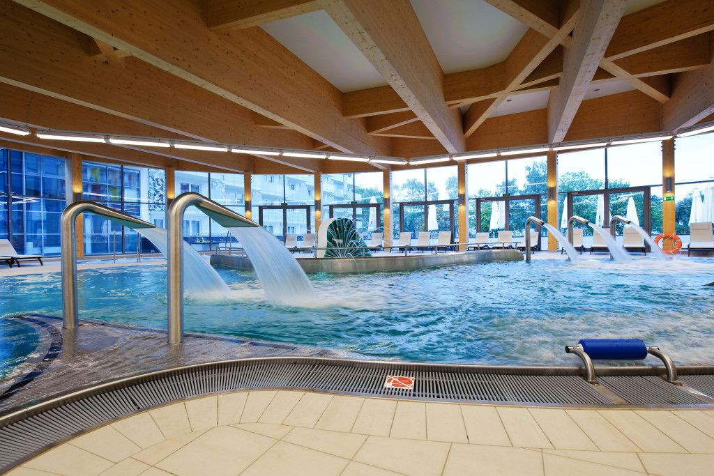 Ein großes Hallenbad mit mehreren Wasserrutschen, gefliestem Boden und Blick nach draußen durch große Fenster.