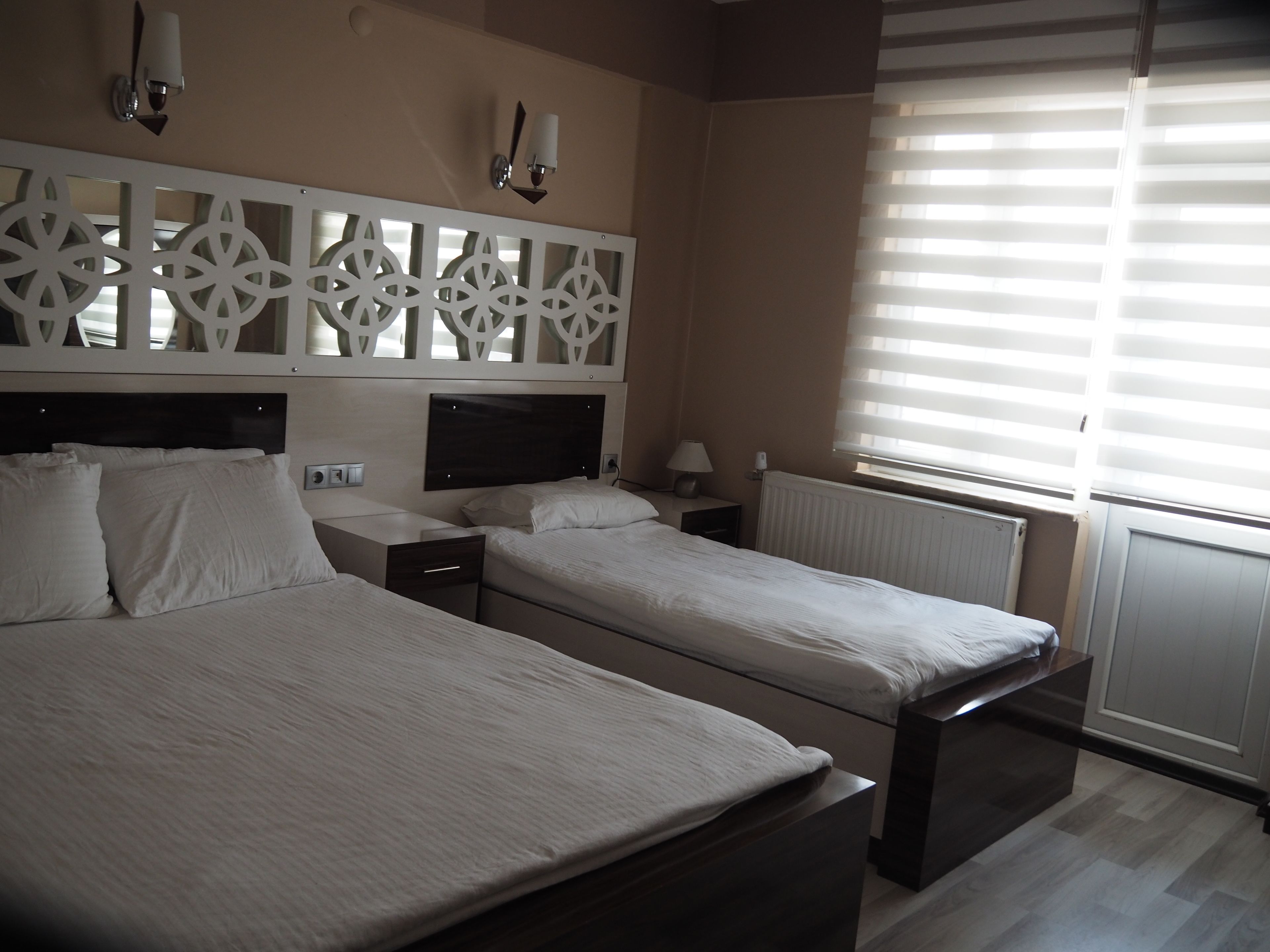 A hotel room with two beds, a decorative wall panel, a bedside table with a lamp, and a window with blinds.