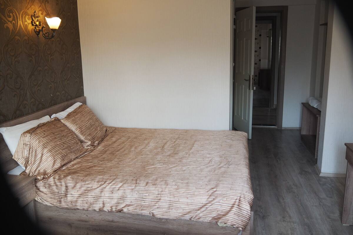 A bedroom with a bed, a nightstand, a wall-mounted light fixture, and a door leading to another room.